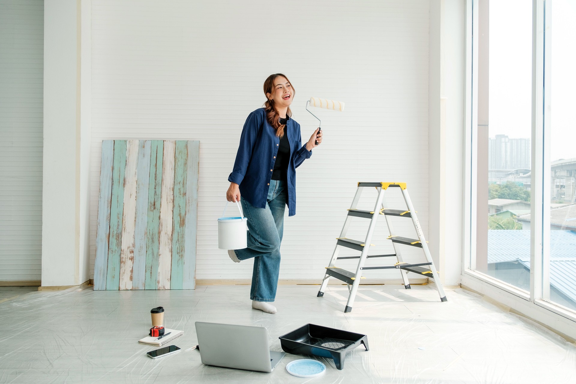 Smiling young woman holding a paint bucket and roller, standing in a bright room during a DIY home makeover. Surrounded by tools and laptop, expressing joy and creativity in home renovation.